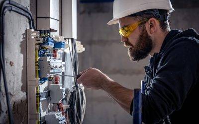 A male electrician works in a switchboard with an electrical connecting cable, connects the equipment with tools.